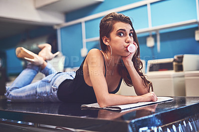 Buy stock photo Portrait of an attractive young woman lying on a counter while blowing bubbles with chewing gum to pass the time in a laundry room