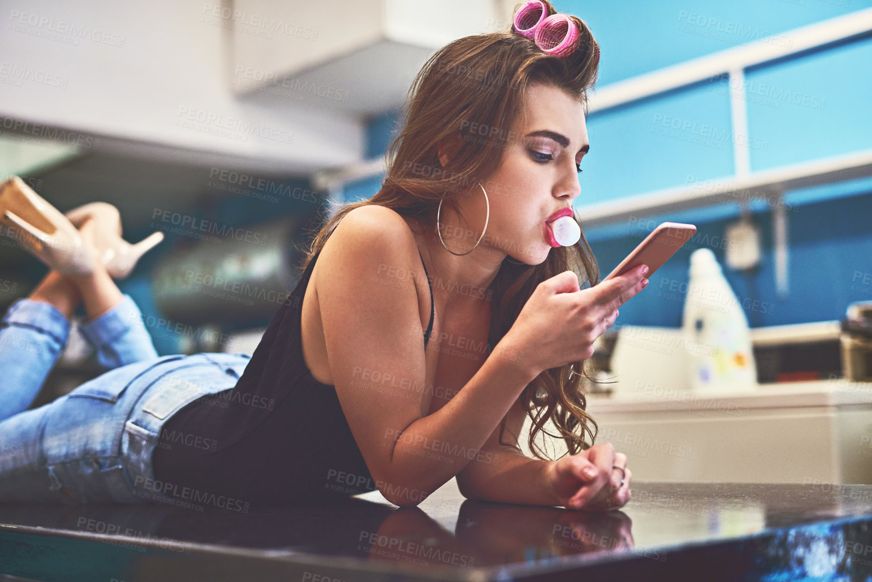 Buy stock photo Shot of an attractive young woman lying on top of a counter while browsing on a cellphone to pass time in a laundry room