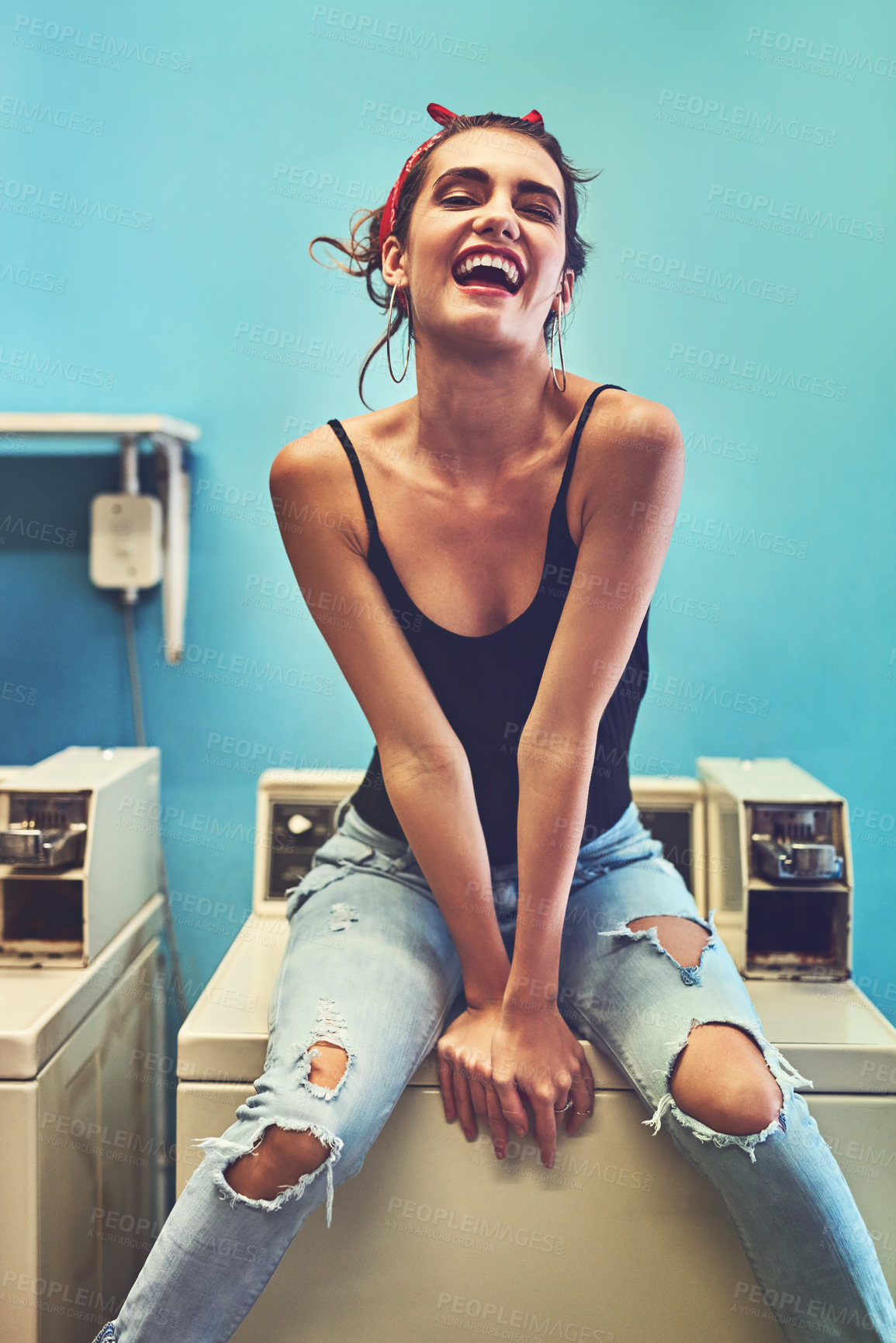Buy stock photo Portrait of an attractive young woman seated on a washing machine while waiting for the washing to be washed inside of a laundry room