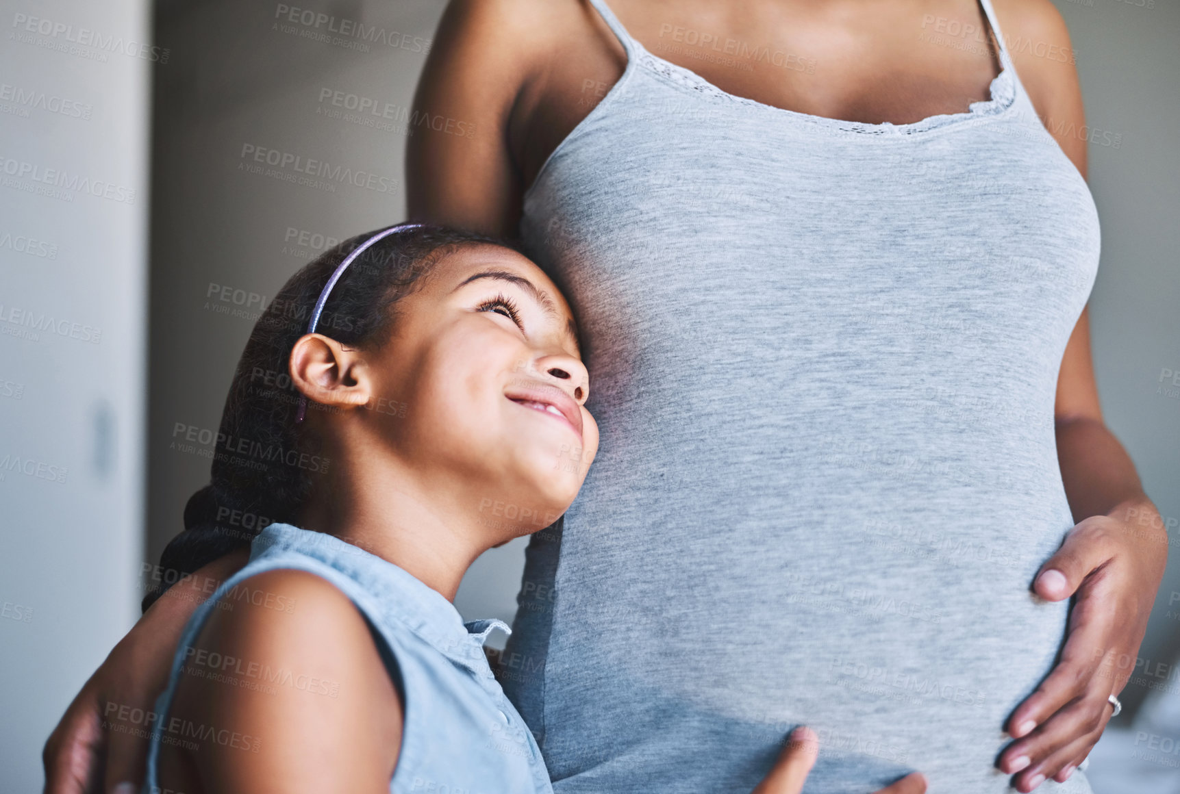 Buy stock photo Shot of a cheerful little girl standing with her arms around her pregnant other at home during the day