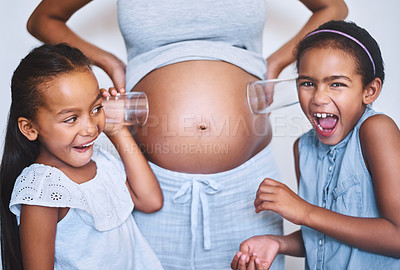 Buy stock photo Portrait of two cheerful little girls standing next their mother while each putting a glass on her pregnant belly to listen for movement at home