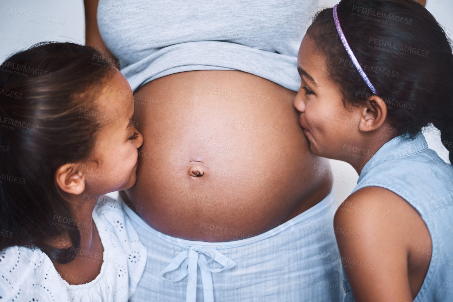 Buy stock photo Shot of two cheerful little girls standing next to their mother and giving her pregnant belly a kiss at home during the day