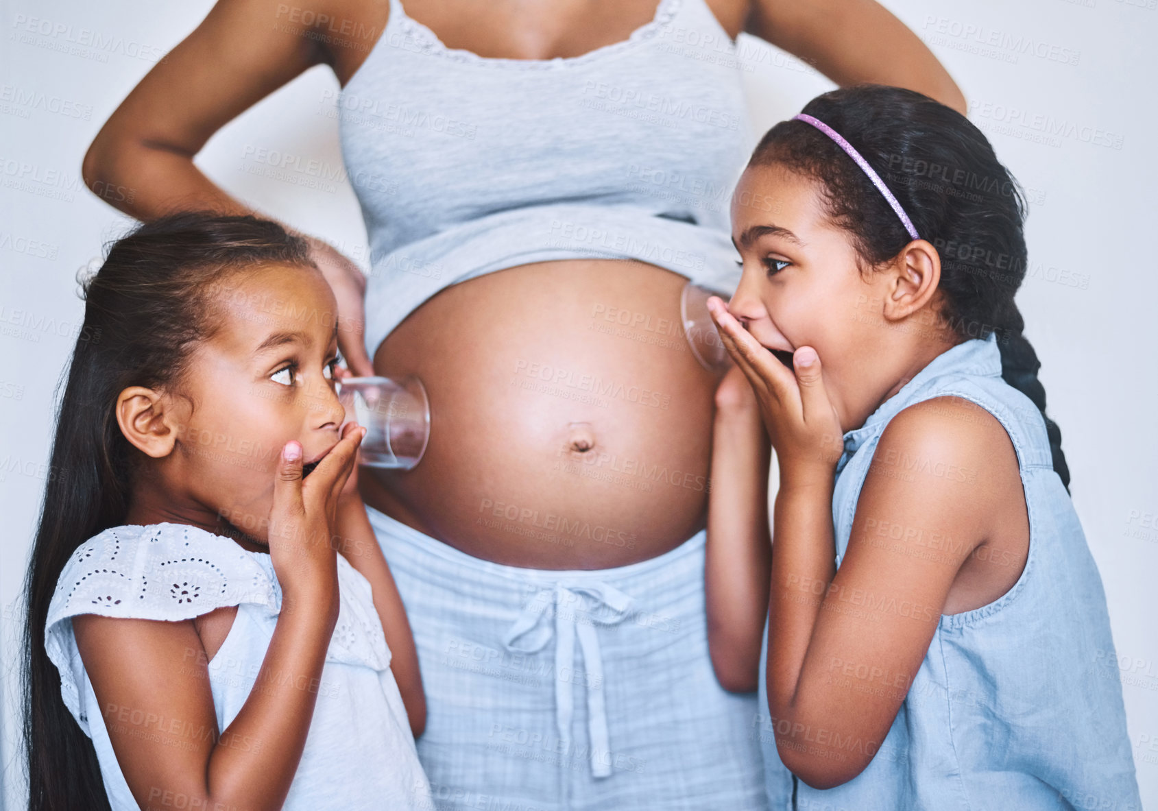 Buy stock photo Shot of two cheerful little girls standing next their mother while each putting a glass on her pregnant belly to listen for movement at home