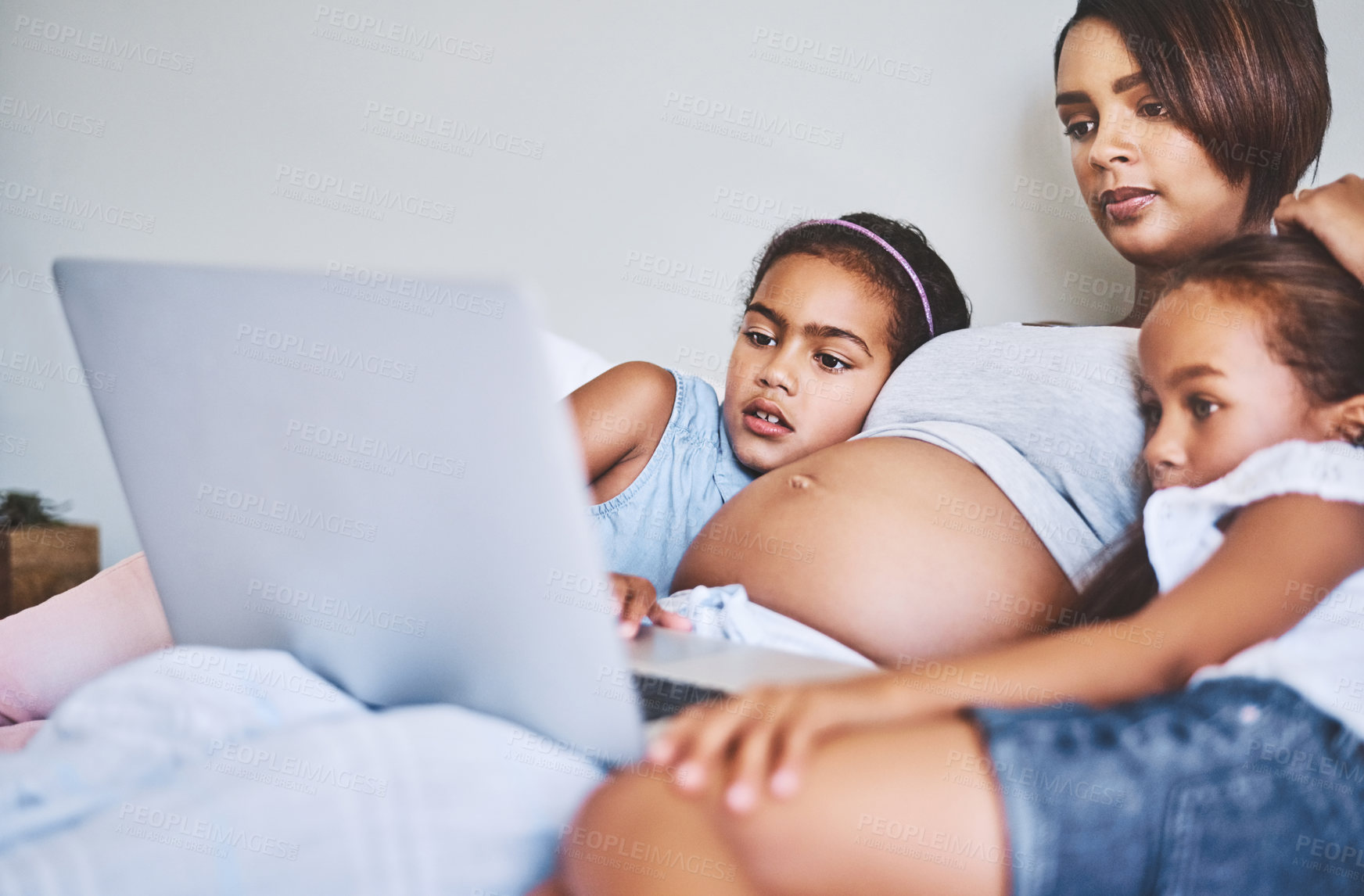 Buy stock photo Shot of two focused little girls relaxing next to their pregnant mother while watching a movie on a laptop at home during the day