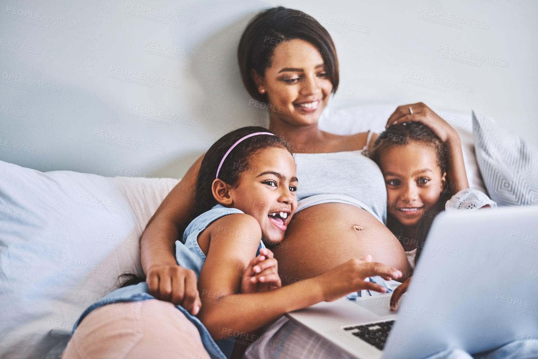 Buy stock photo Shot of two cheerful little girls relaxing next to their pregnant mother while watching a movie on a laptop at home during the day