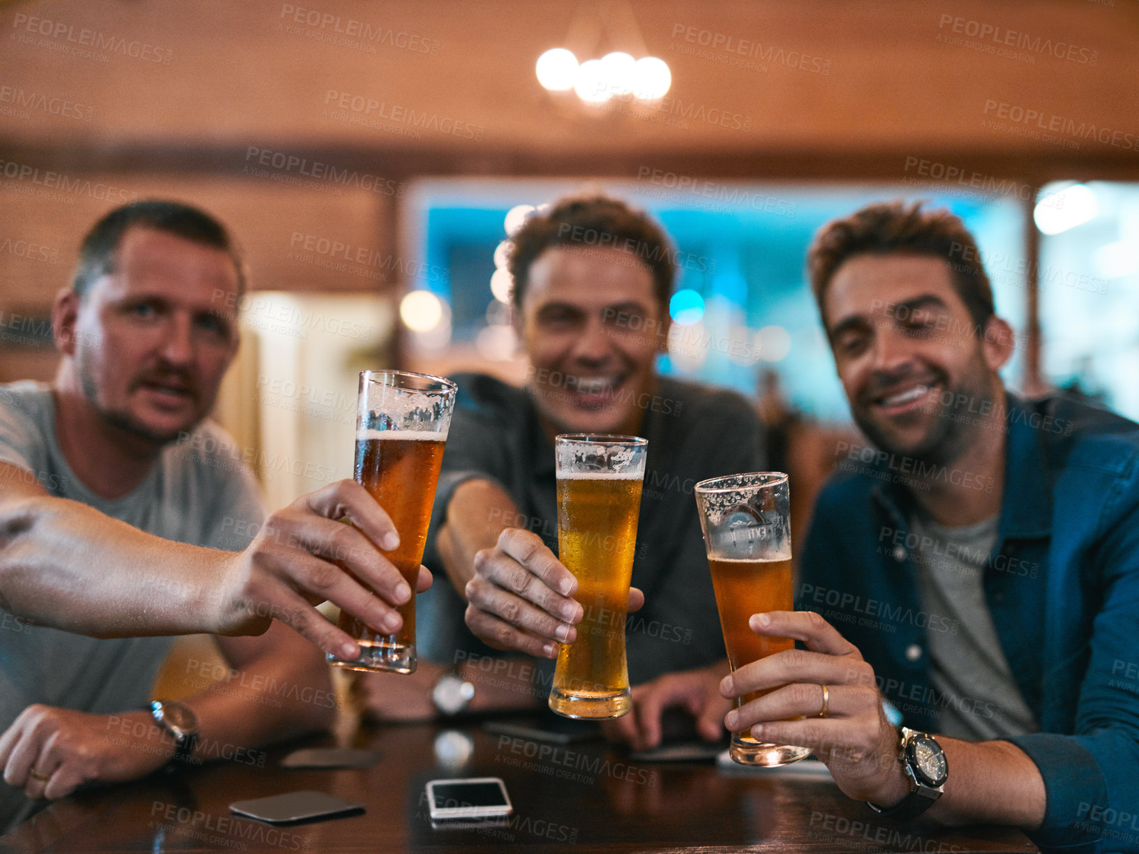Buy stock photo Portrait of three cheerful young men having a celebratory toast with beer while sitting at a table inside of a beer brewery during the day