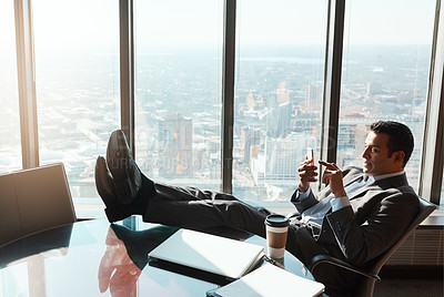 Buy stock photo Shot of a young businessman using a cellphone while relaxing with his feet up on a table in an office