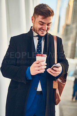 Buy stock photo Shot of a cheerful young man texting on his phone while drinking coffee in the busy streets of the city while going to work in the morning