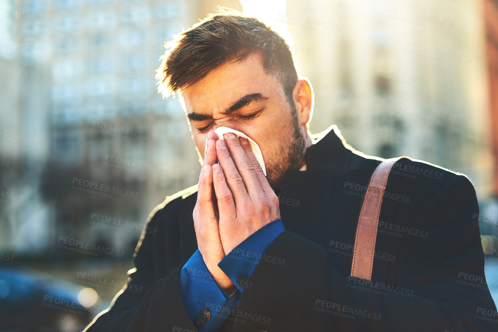Buy stock photo Shot of a irritated looking young man blowing his nose with a tissue while walking the busy streets of the city in the morning
