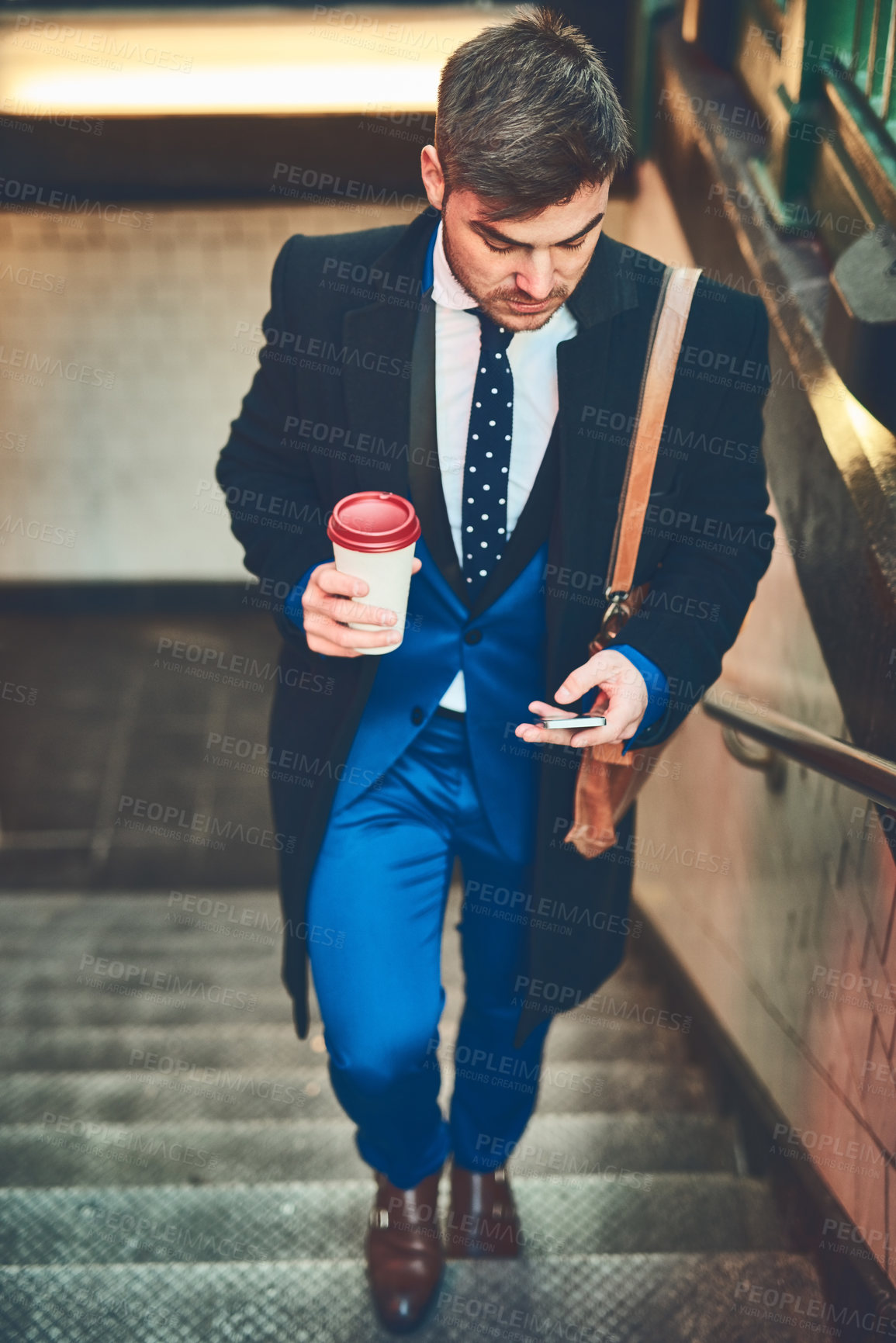 Buy stock photo Shot of a focused young businessman texting on his phone and drinking coffee while walking up a flight of stairs in a subway during the morning hours