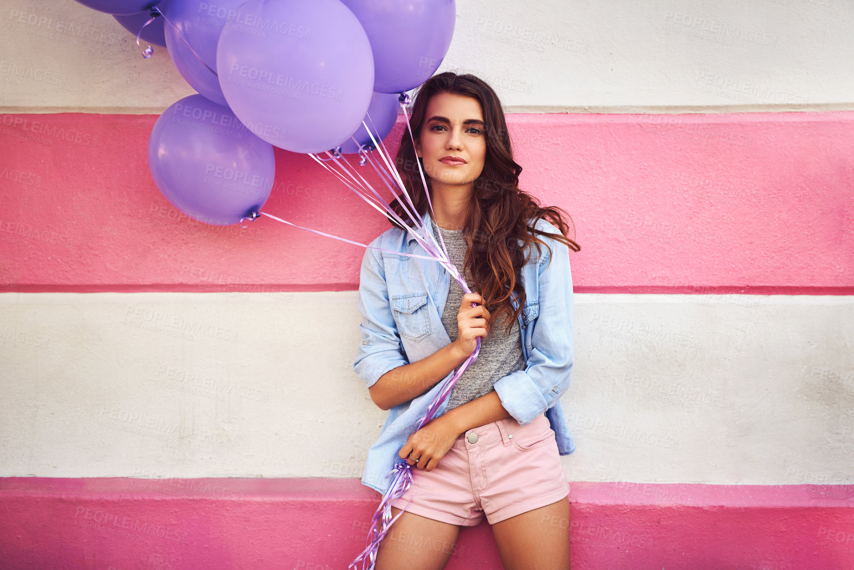 Buy stock photo Cropped shot of a beautiful young woman holding purple balloons against a wall outside