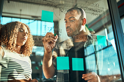 Buy stock photo Shot of two focussed young work colleagues putting up sticky notes on a glass wall while brainstorming together in the office at work