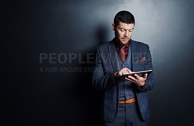 Buy stock photo Cropped shot of a handsome young businessman using his tablet while standing against a dark background