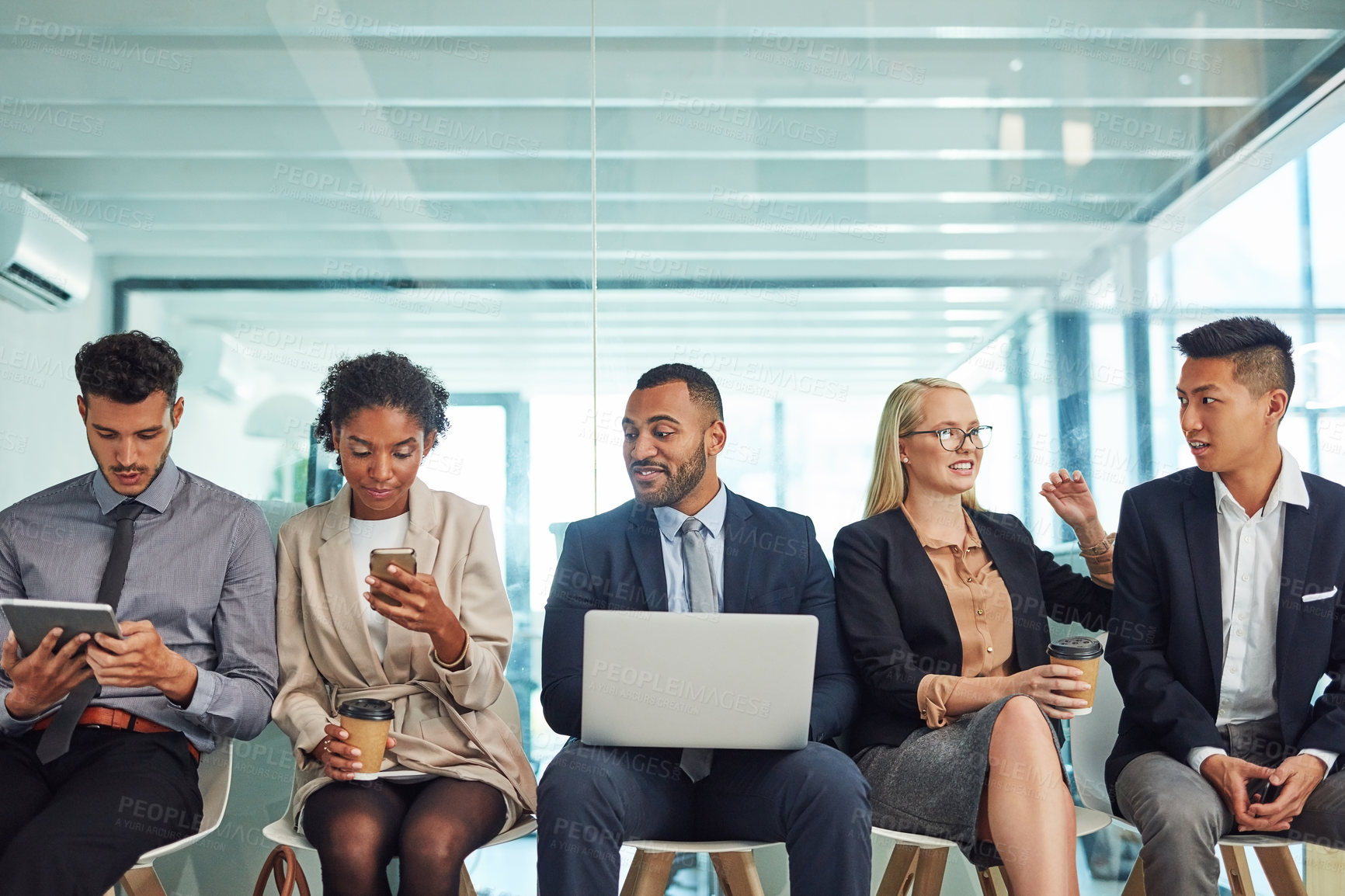 Buy stock photo Shot of a group of focussed young businesspeople seated together on chairs and making notes using different methods while waiting in the office during the day
