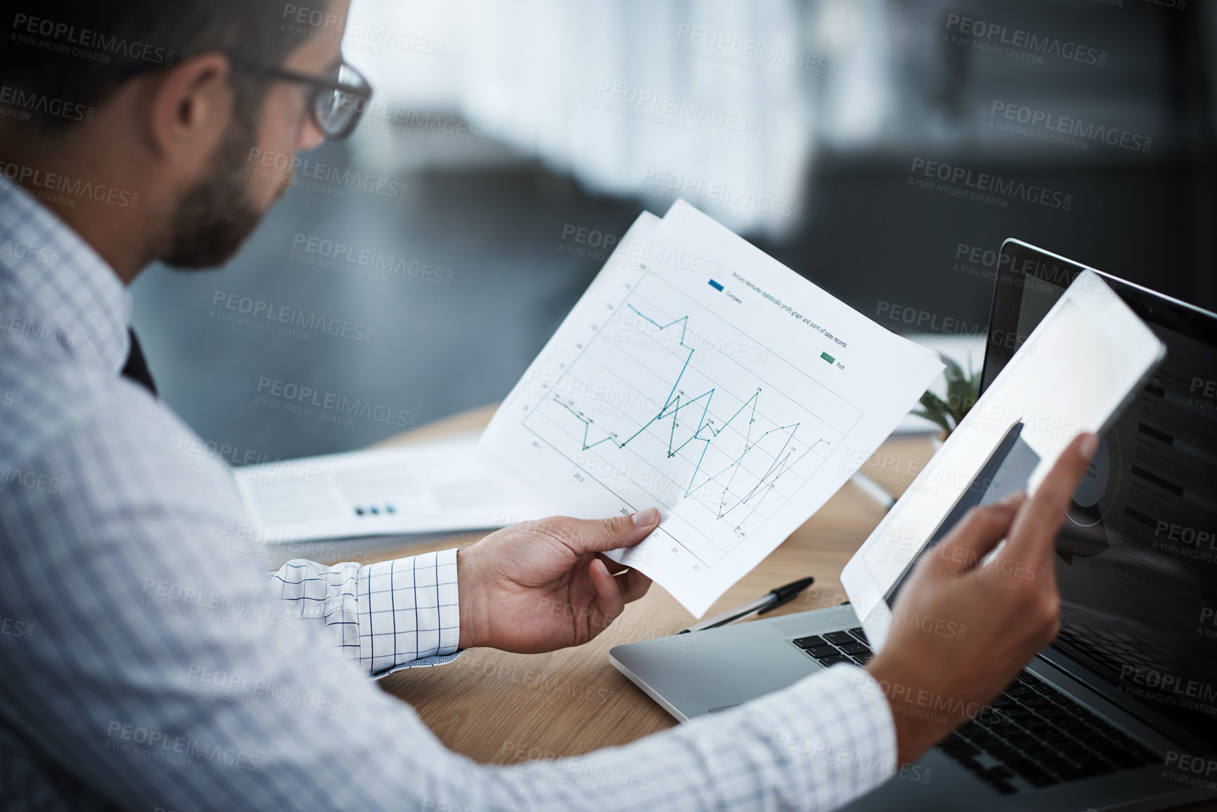 Buy stock photo Shot of a young businessman using a digital tablet while analyzing graphs in an office
