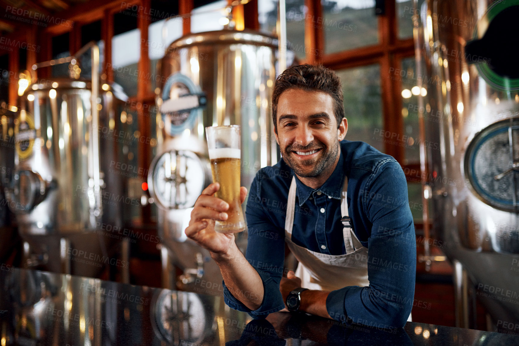 Buy stock photo Portrait of a cheerful young business owner holding up a glass of beer that he just poured inside of a beer brewery during the day