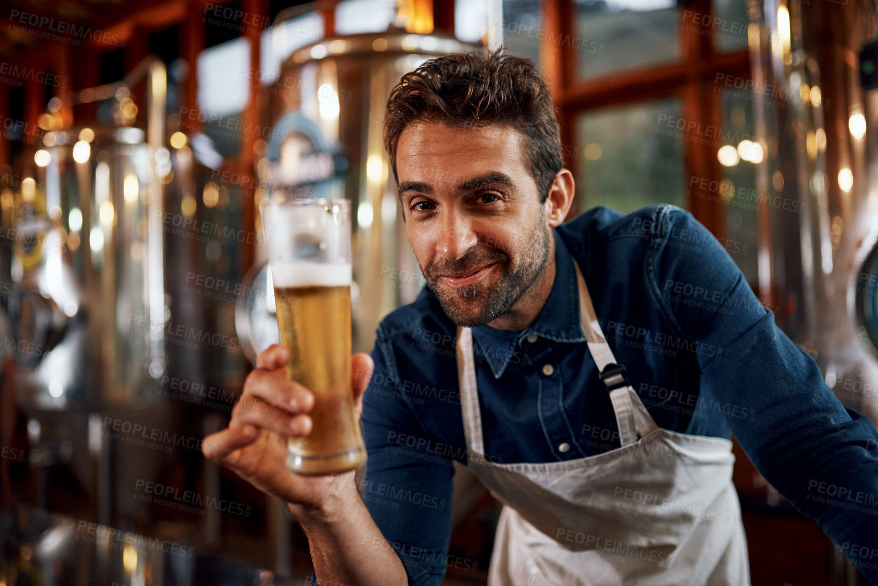 Buy stock photo Portrait of a cheerful young business owner holding up a glass of beer that he just poured inside of a beer brewery during the day