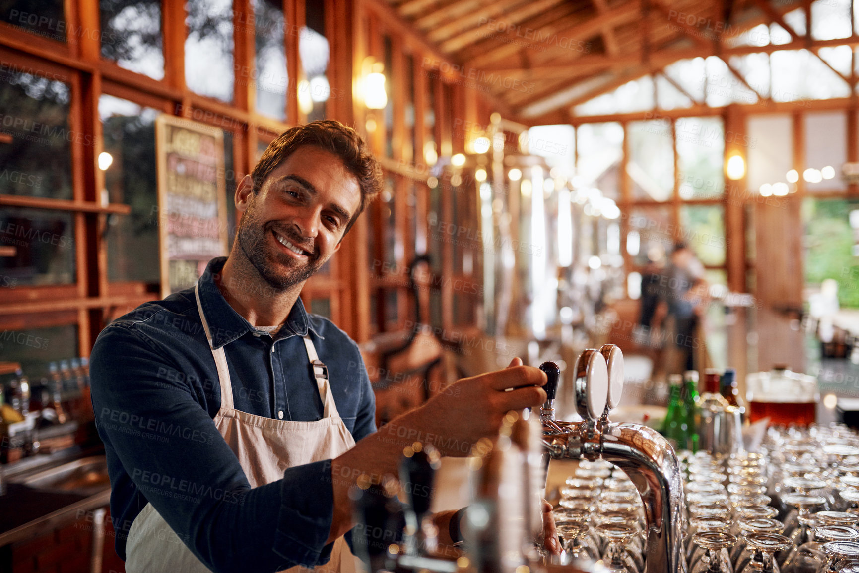 Buy stock photo Portrait of a cheerful young barman pouring a glass of beer inside of a beer brewery during the day