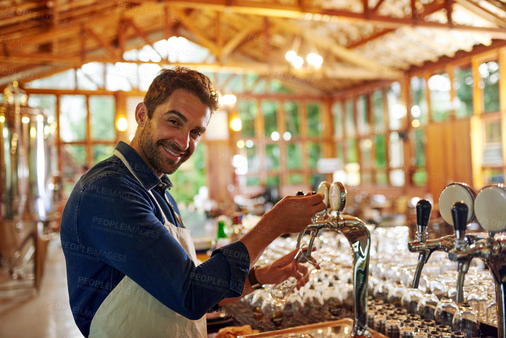 Buy stock photo Portrait of a cheerful young barman pouring a glass of beer inside of a beer brewery during the day