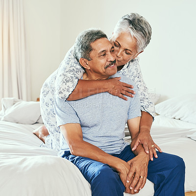 Buy stock photo Shot of a cheerful mature man being held by his wife and receiving a kiss on the cheek while they both sit on the bed at home