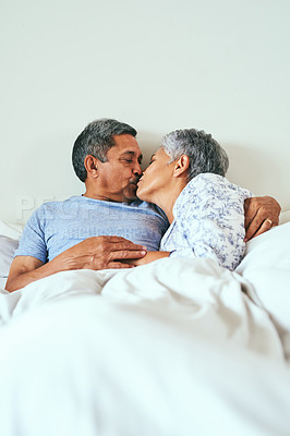 Buy stock photo Shot of a cheerful mature couple relaxing in bed while sharing a kiss at home during the day