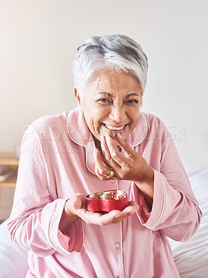 Buy stock photo Portrait of a cheerful mature woman enjoying a piece of chocolate while being seated on a bed at home in the morning