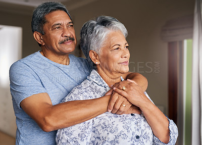 Buy stock photo Shot of a cheerful mature couple holding each other while looking outside through a window at home during the day