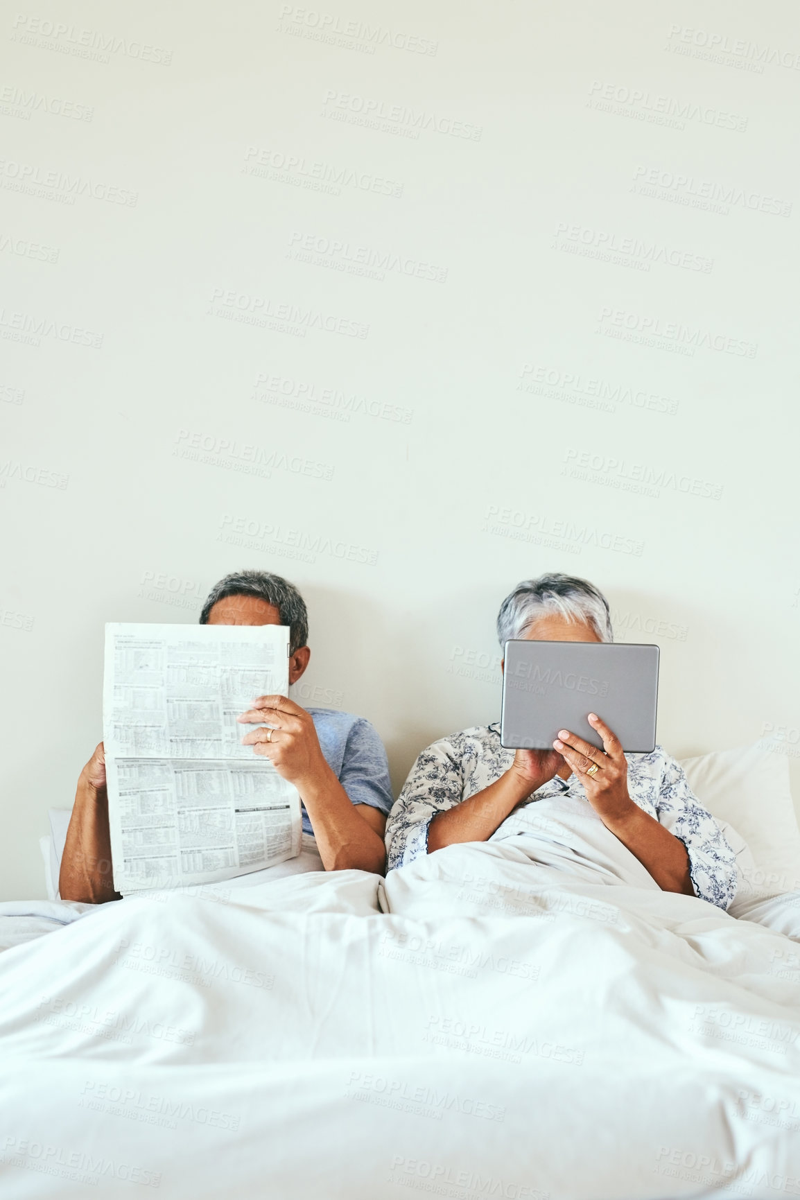 Buy stock photo Shot of a relaxed mature couple lying in bed while browsing on a digital tablet and reading the newspaper at home in the morning