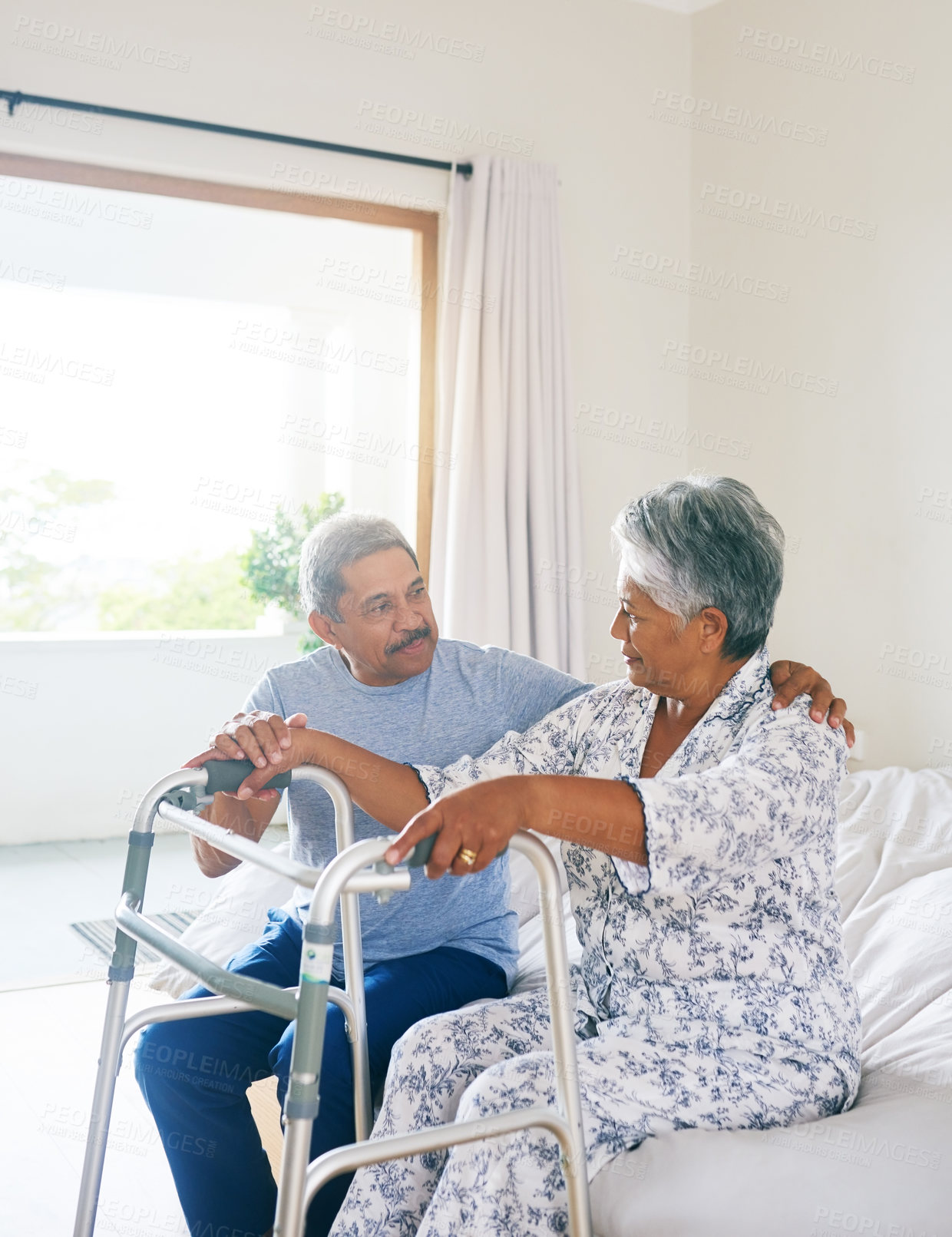 Buy stock photo Shot of a determined mature woman seated on a bed and about to walk with a walking frame while her husband supports her at home