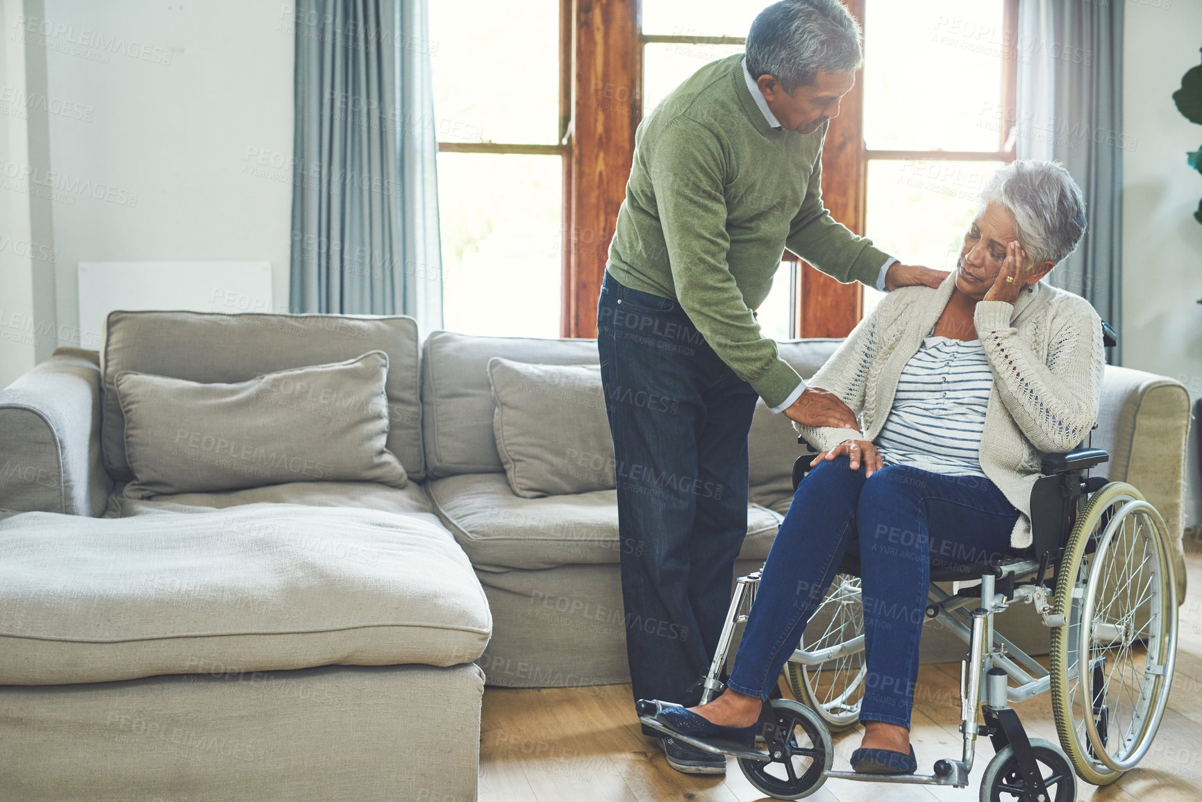 Buy stock photo Shot of a worried looking elderly woman seated in a wheelchair while being supported and held by her husband inside at home during the day