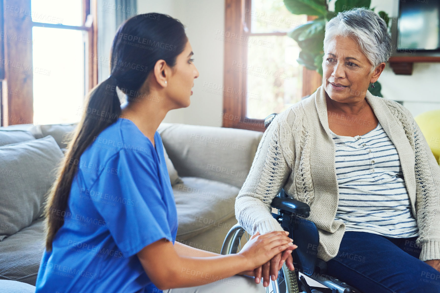 Buy stock photo Shot of a worried looking elderly woman seated in a wheelchair while a female nurse holds her hand for support inside at home during the day