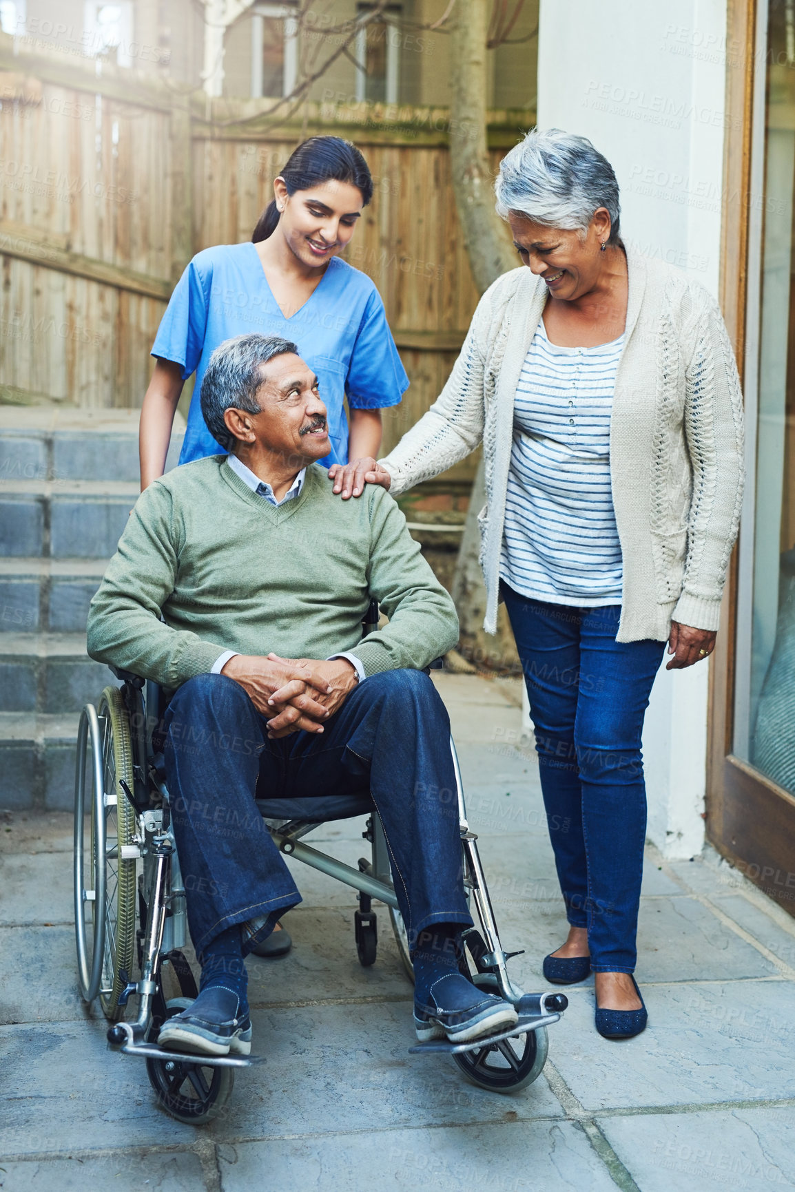 Buy stock photo Shot of a cheerful elderly man seated in a wheelchair while being supported by his wife and a female nurse at home during the day