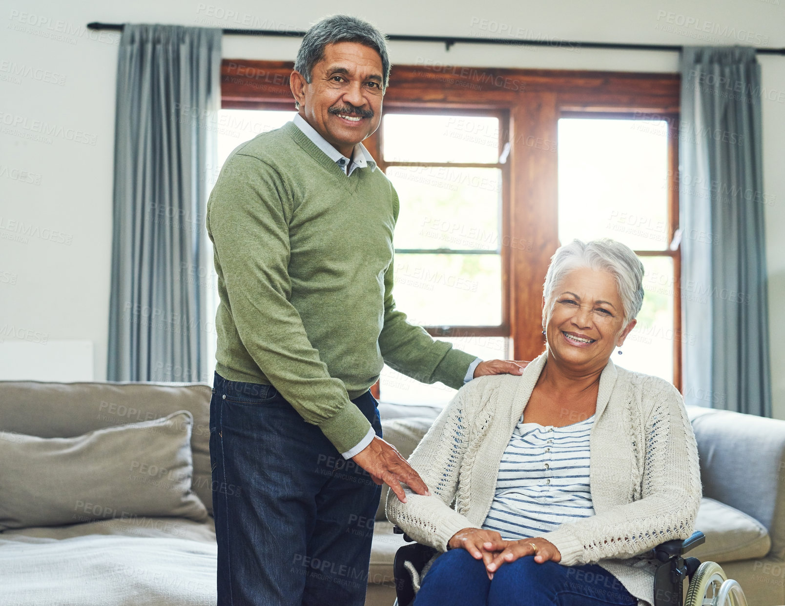 Buy stock photo Portrait of a cheerful elderly woman seated in a wheelchair while being supported and held by her husband inside at home during the day