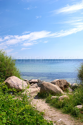 Buy stock photo A path to the shore with rocks and lush green grass. Scenic view of holiday destination. A calm summer day at the beach outdoors in nature. Landscape view of the sea in spring with a cloudy blue sky