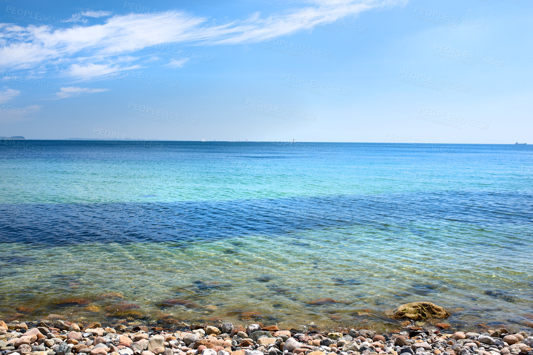 Buy stock photo Calm ocean waves washing onto stones at an empty beach shore with sailboats cruising in the horizon. Scenic landscape for a relaxing summer vacation. Copy space at the sea with a blue sky background