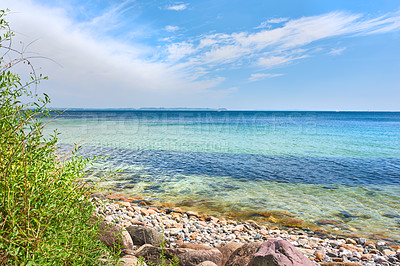 Buy stock photo Copyspace at sea with a cloudy blue sky background. Calm ocean waves washing onto stones at an empty beach shore with sailboats cruising in the horizon. Scenic landscape for a relaxing summer holiday