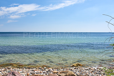 Buy stock photo Copy space at the sea with a blue sky background. Calm ocean waves washing onto stones at an empty beach shore with sailboats cruising in the horizon. Scenic landscape for a relaxing summer holiday