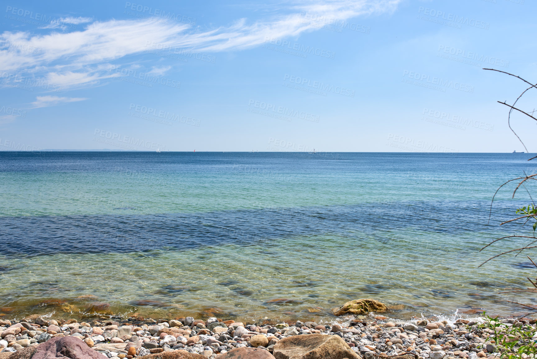 Buy stock photo Copy space at the sea with a blue sky background. Calm ocean waves washing onto stones at an empty beach shore with sailboats cruising in the horizon. Scenic landscape for a relaxing summer holiday