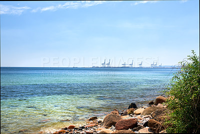 Buy stock photo Copy space at sea with a blue sky background and harbour cranes at a port in the horizon. Calm ocean waves at a shallow and rocky shore. Scenic coastal landscape for a relaxing summer holiday