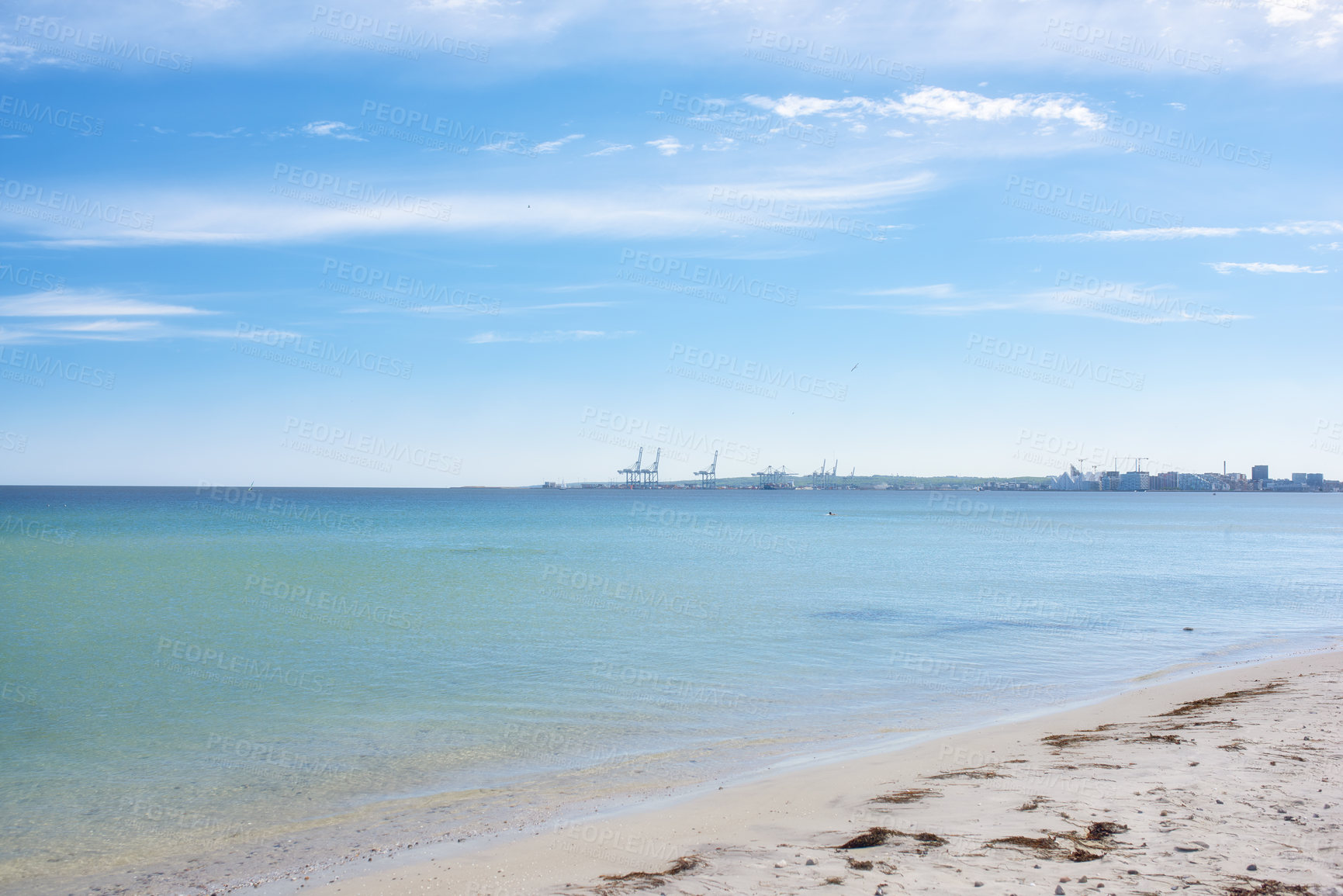 Buy stock photo Tranquil ocean waves wash onto the shore at an empty beach with a port or harbour on the horizon. A scenic landscape for a relaxing summer holiday or vacation. Calm sea under blue sky copy space.