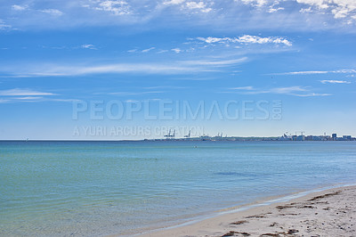 Buy stock photo Copy space at the sea with a blue sky background. Calm ocean waves wash onto stones at an empty beach shore with a port or harbor on the horizon. a scenic landscape for a relaxing summer holiday