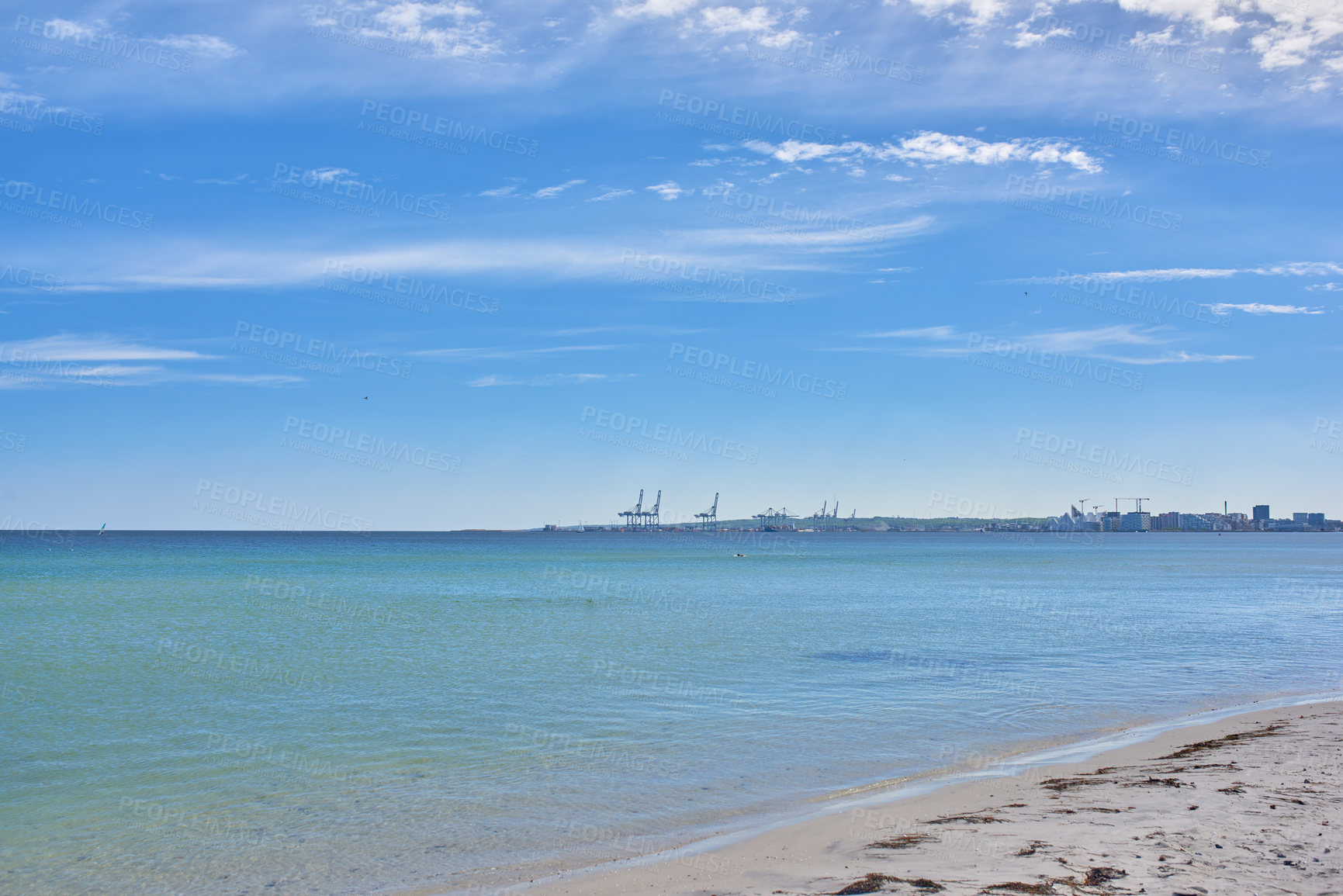 Buy stock photo Copy space at the sea with a blue sky background. Calm ocean waves wash onto stones at an empty beach shore with a port or harbor on the horizon. a scenic landscape for a relaxing summer holiday