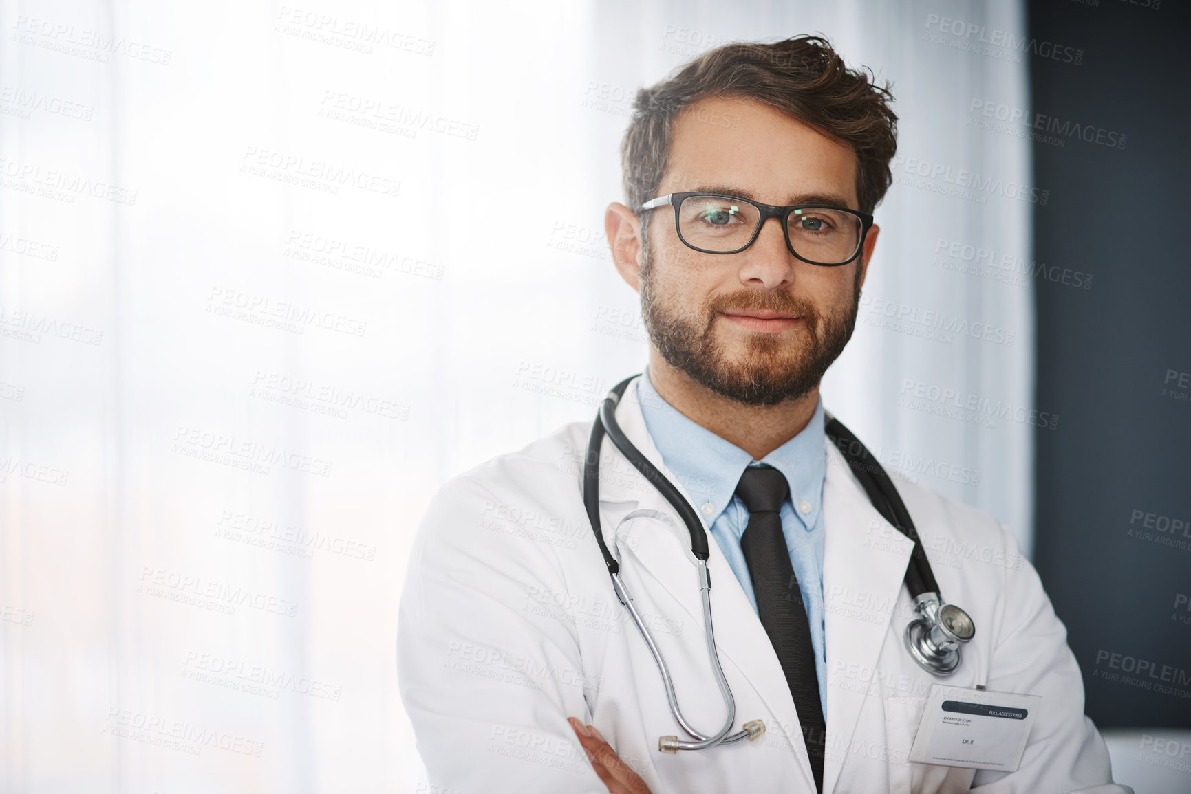 Buy stock photo Cropped portrait of a confident young male doctor standing with his arms folded inside of a hospital