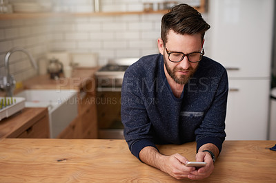 Buy stock photo Cropped shot of a handsome young man sending a text message while standing in the kitchen at home
