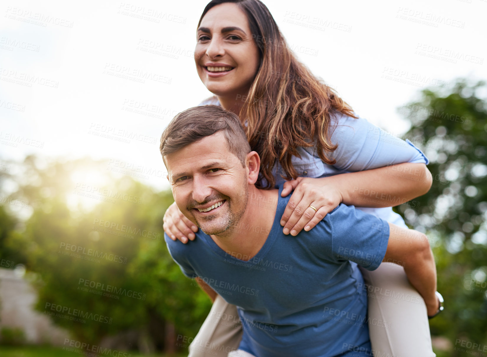 Buy stock photo Shot of a cheerful young man giving his wife a piggyback ride outside in a park during the day