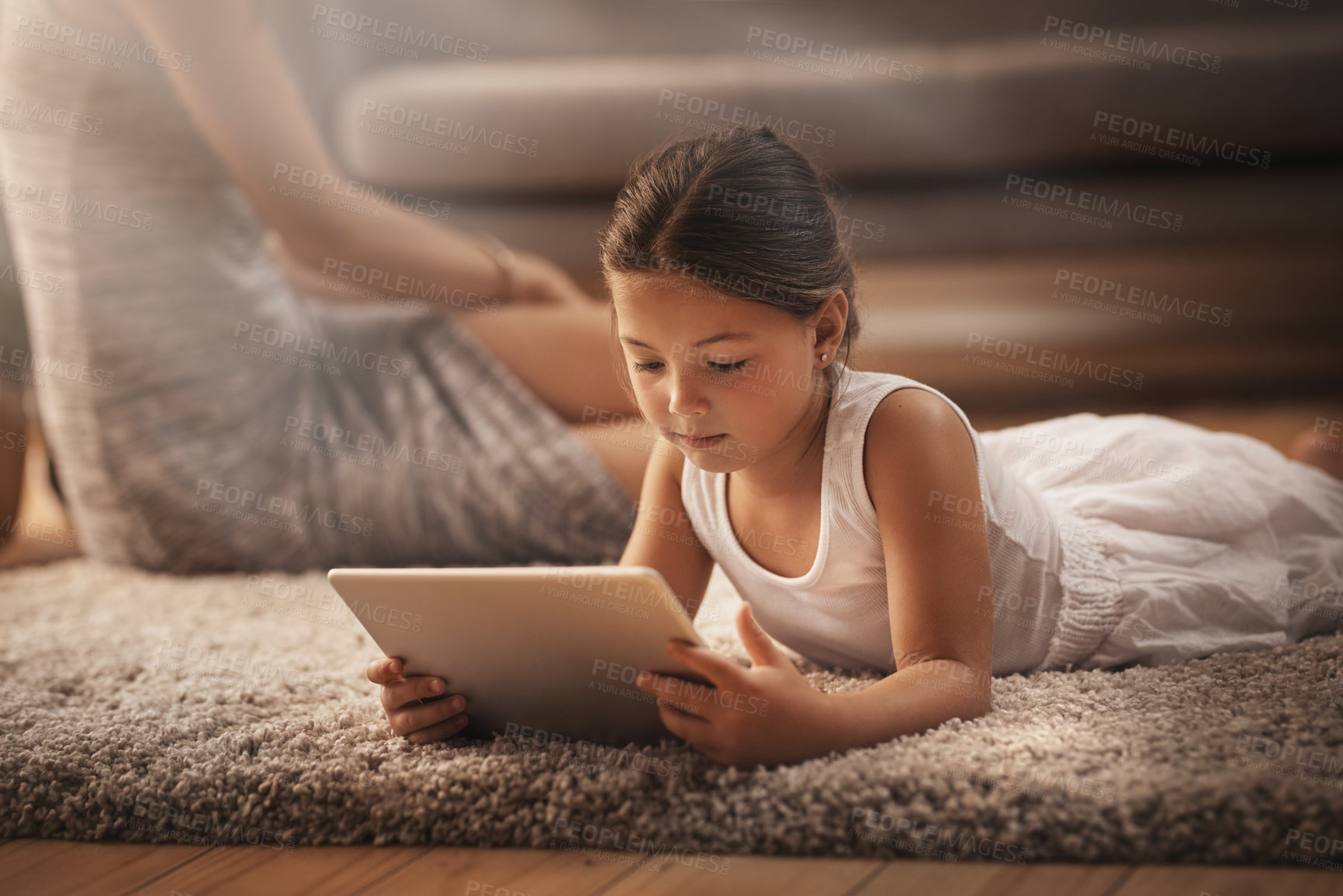 Buy stock photo Shot of an adorable little girl using a digital tablet on the floor at home with her mother in the background