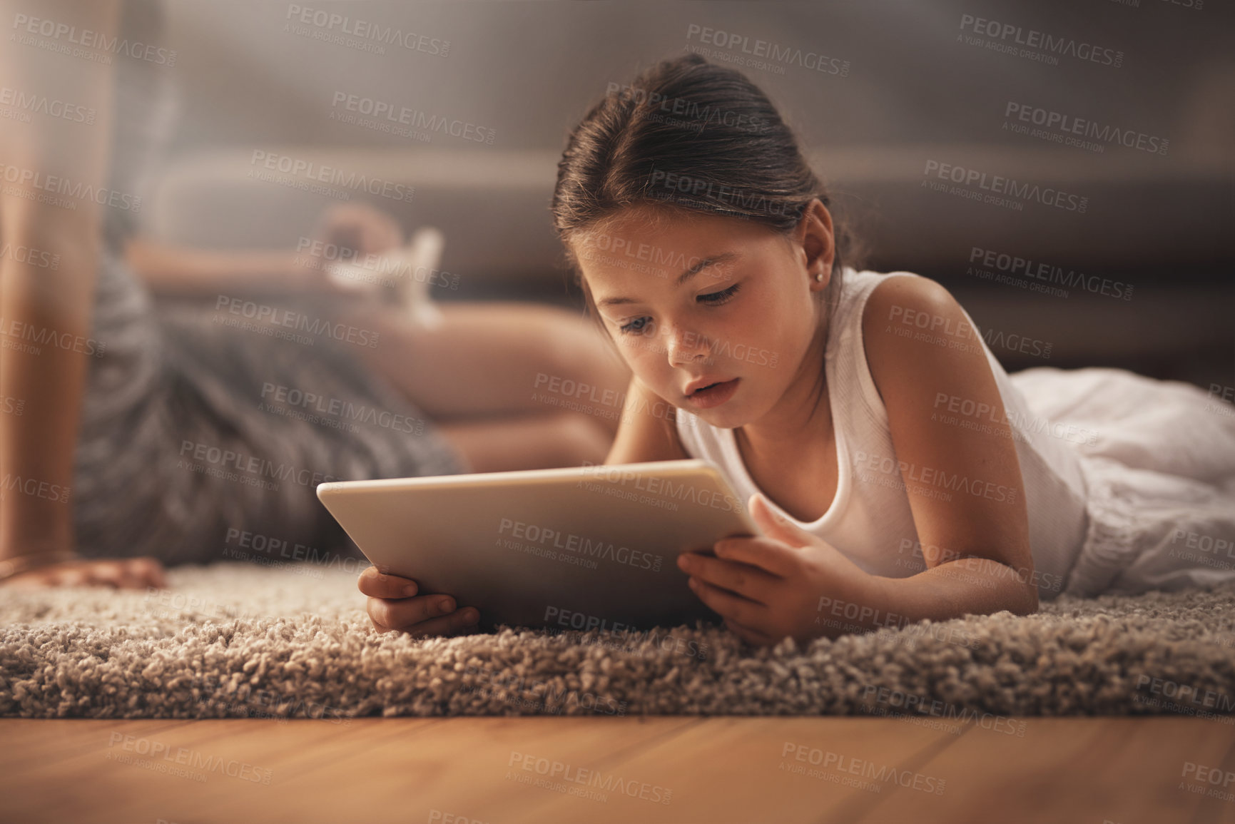 Buy stock photo Shot of an adorable little girl using a digital tablet on the floor at home with her mother in the background