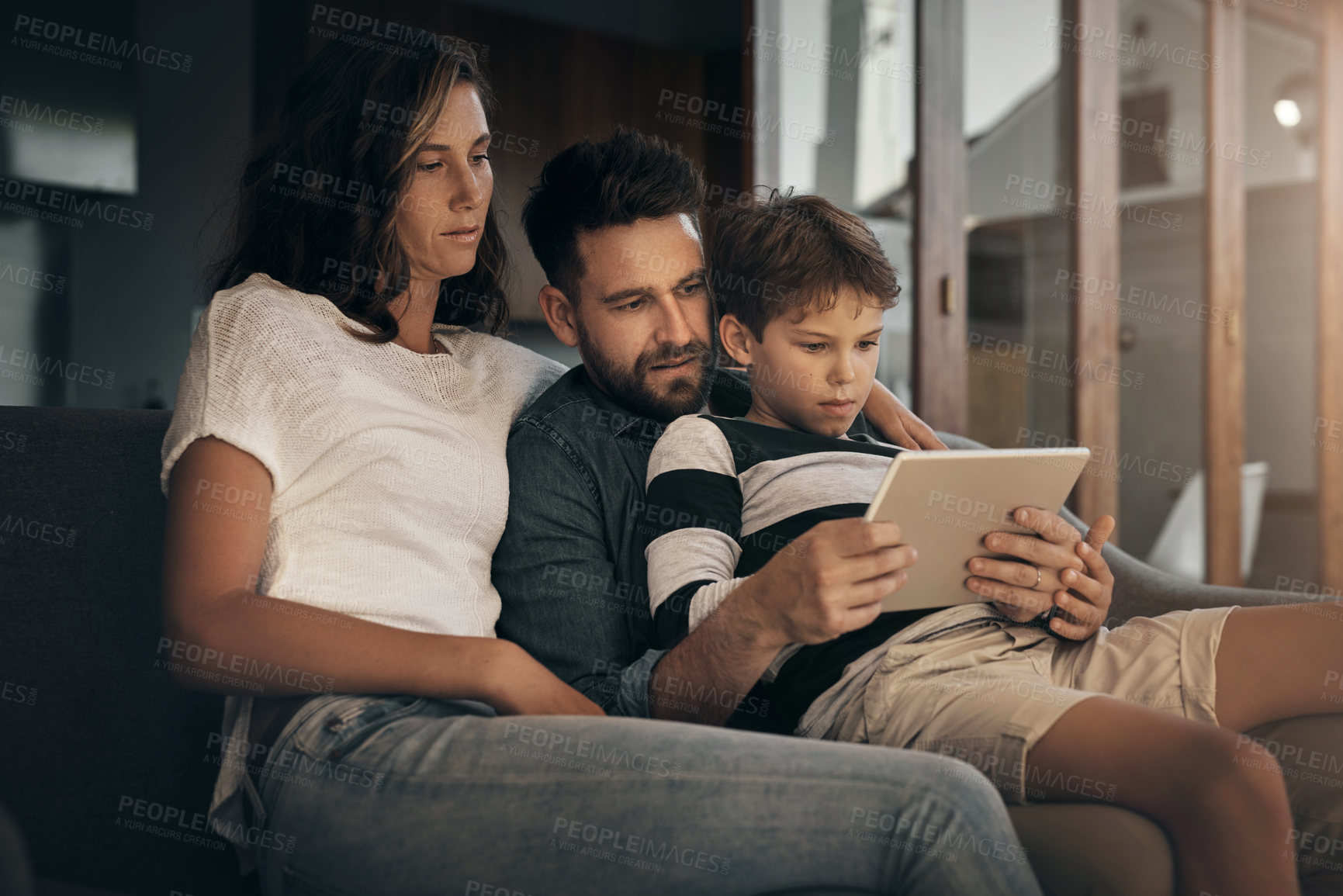 Buy stock photo Shot of an adorable little boy using a digital tablet with his mother and father on the sofa at home