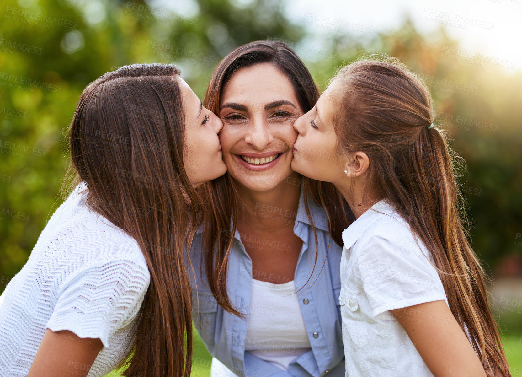 Buy stock photo Portrait of a cheerful young woman receiving a kiss on each cheek from her young daughters outside in a park during the day