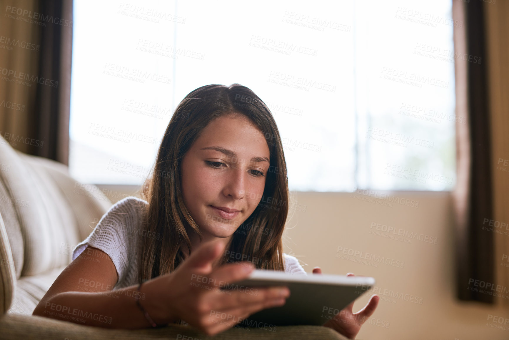 Buy stock photo Shot of a focussed young girl browsing on a digital tablet while lying on a sofa at home during the day